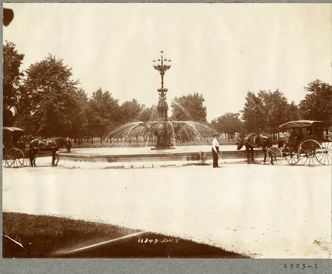 Black and white photograph of dirt center surrounded by grass and trees with water fountain at center. Horses dip down to drink water in the fountain