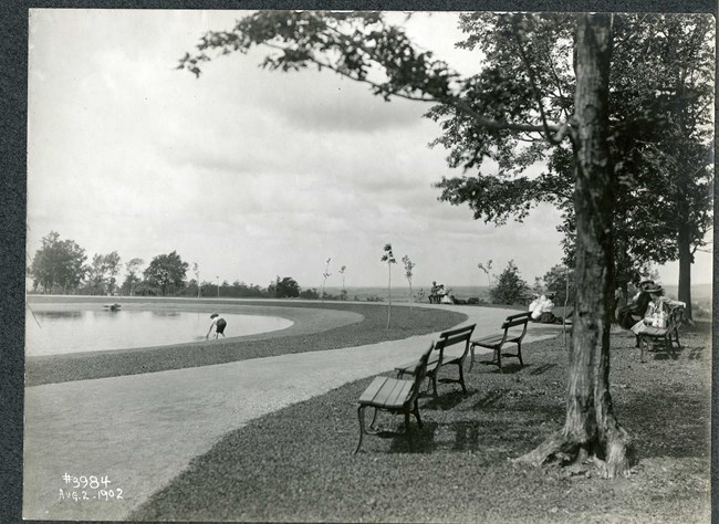 Black and white of flat top of hill with body of water on top. There is a path around the water with some trees. benches and people. There is a person leaned over the water.