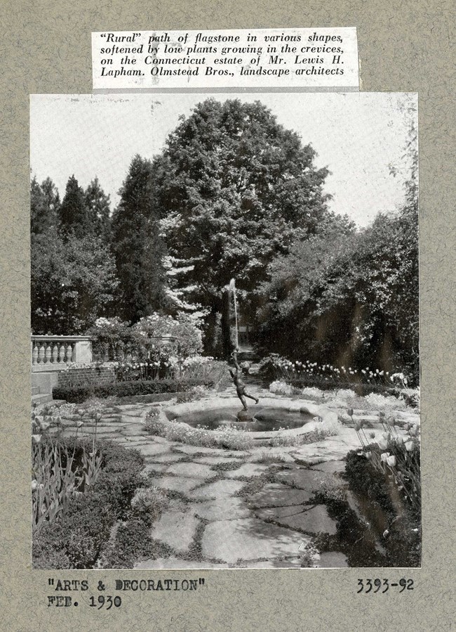 Black and white photograph of garden, with stone walkways leading to a central area with a circular fountain and statue on top. The area around the garden is planted densely with trees and shrubs.