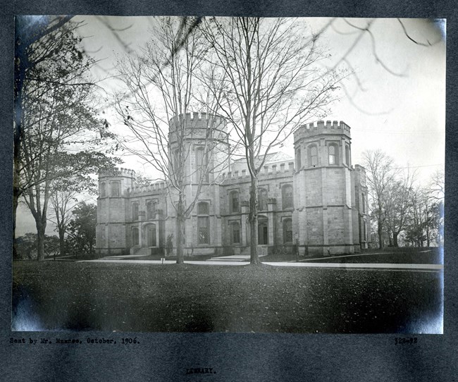 Black and white of flat grassy area with trees lining a path and a large castle-like building.