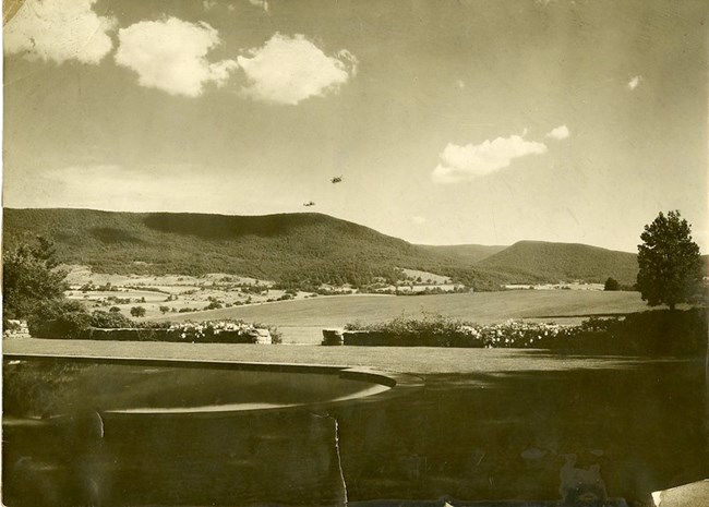 Black and white photograph of large open field surrounded by hills covered with trees