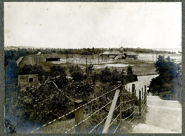 Black and white of large playground area surrounded  by trees with water in the distance.