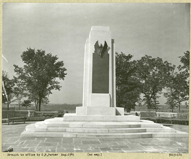 Black and white of stone pillar with wings on it on a hill with trees around the area.