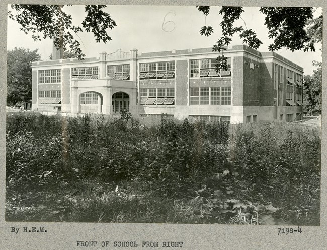 Black and white of school building with dense plantings of shrubs in the front, with trees along the edges of the building.