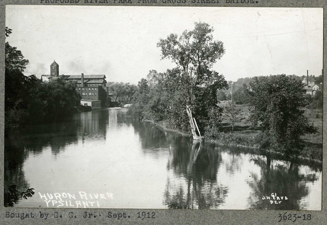 Black and white of body of water with grass and trees on the side, with a large building rising in the distance.
