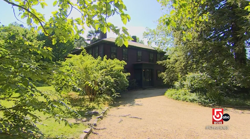 Dirt path leading to large maroon building with grass and trees on one side, large tree in front.