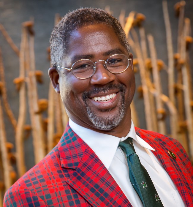 Man smiles wearing red plaid jacket, glasses, and a green tie.