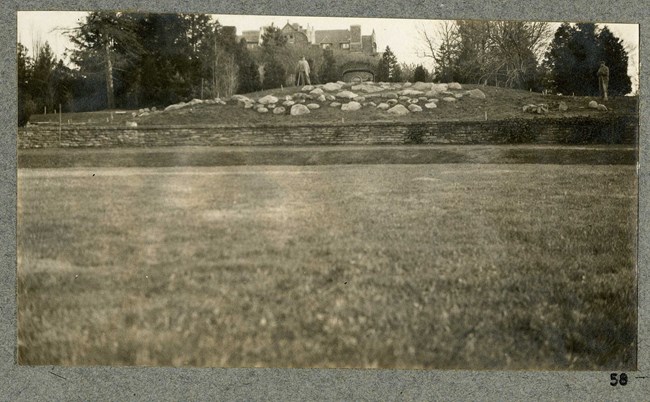 Black and white of flat grassy area leading to a rock wall with rocks on top and a man standing on it and further up the hill are trees and the home.