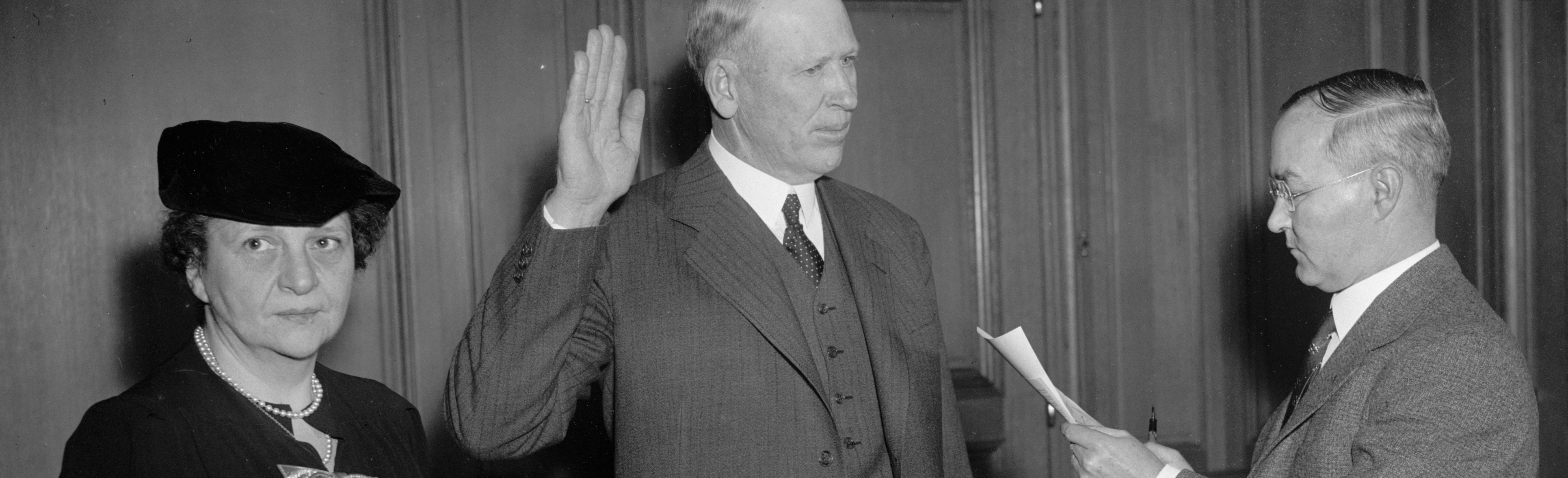 Francis Perkins stands to the left of frame looking at the camera as a man in the center of frame raises his right hand and takes an oath of office.