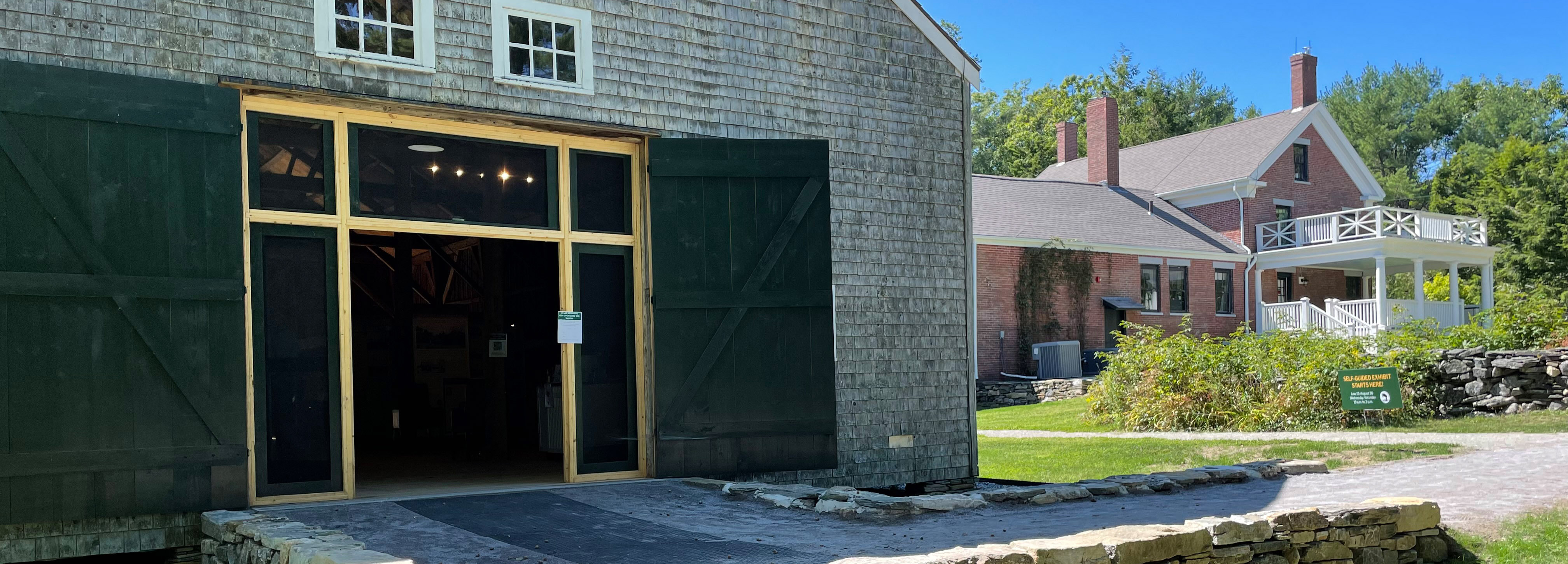 A grey barn with open doors in foreground and a red brick house in the distance below a clear blue sky