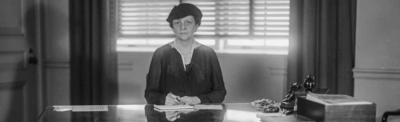 Frances Perkins, seated directly at the center of the frame behind a desk, looking directly at the camear, with a pen in hand as if about to sign the document in front of her.