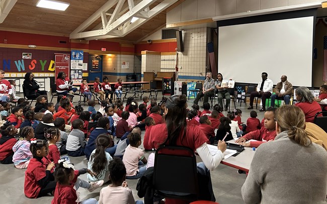 Two ranges reading a book to a group of students in a school lunchroom.