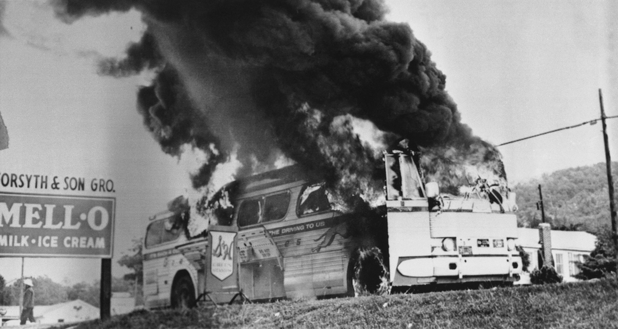 A Greyhound Bus engulfed in flames with smoke pouring out of the door and windshield. A Forsyth and Son Grocery Store is seen next to the sign