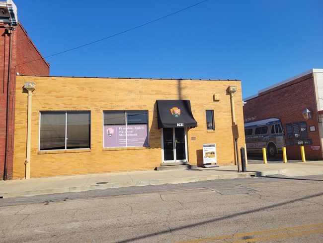 A brick building with a historical marker sits next to the road. A mural of a bus is seen on the next building to the right.