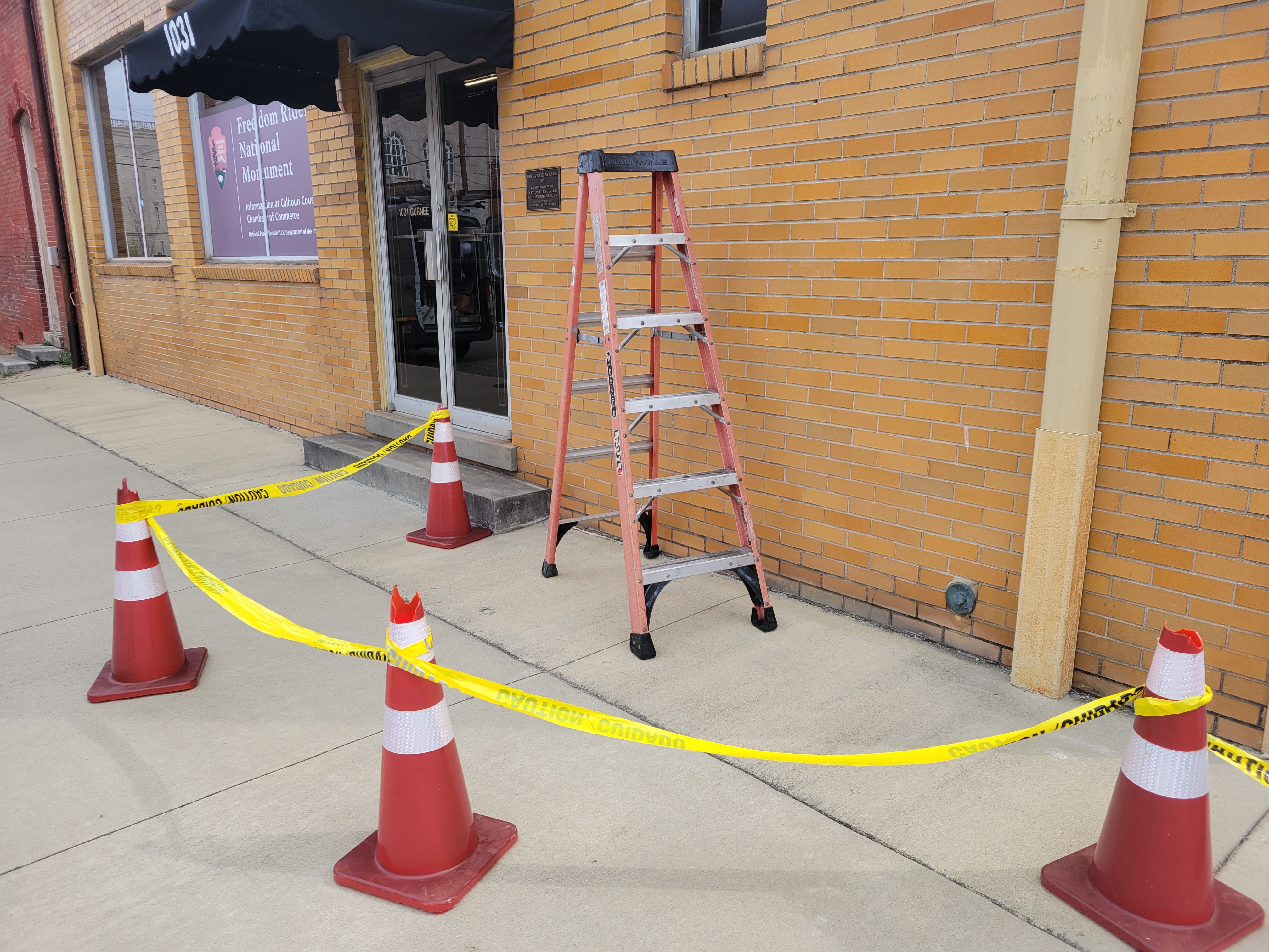 A ladder stands in front of a brick building. A circle of cones and safety tape surround the ladder.