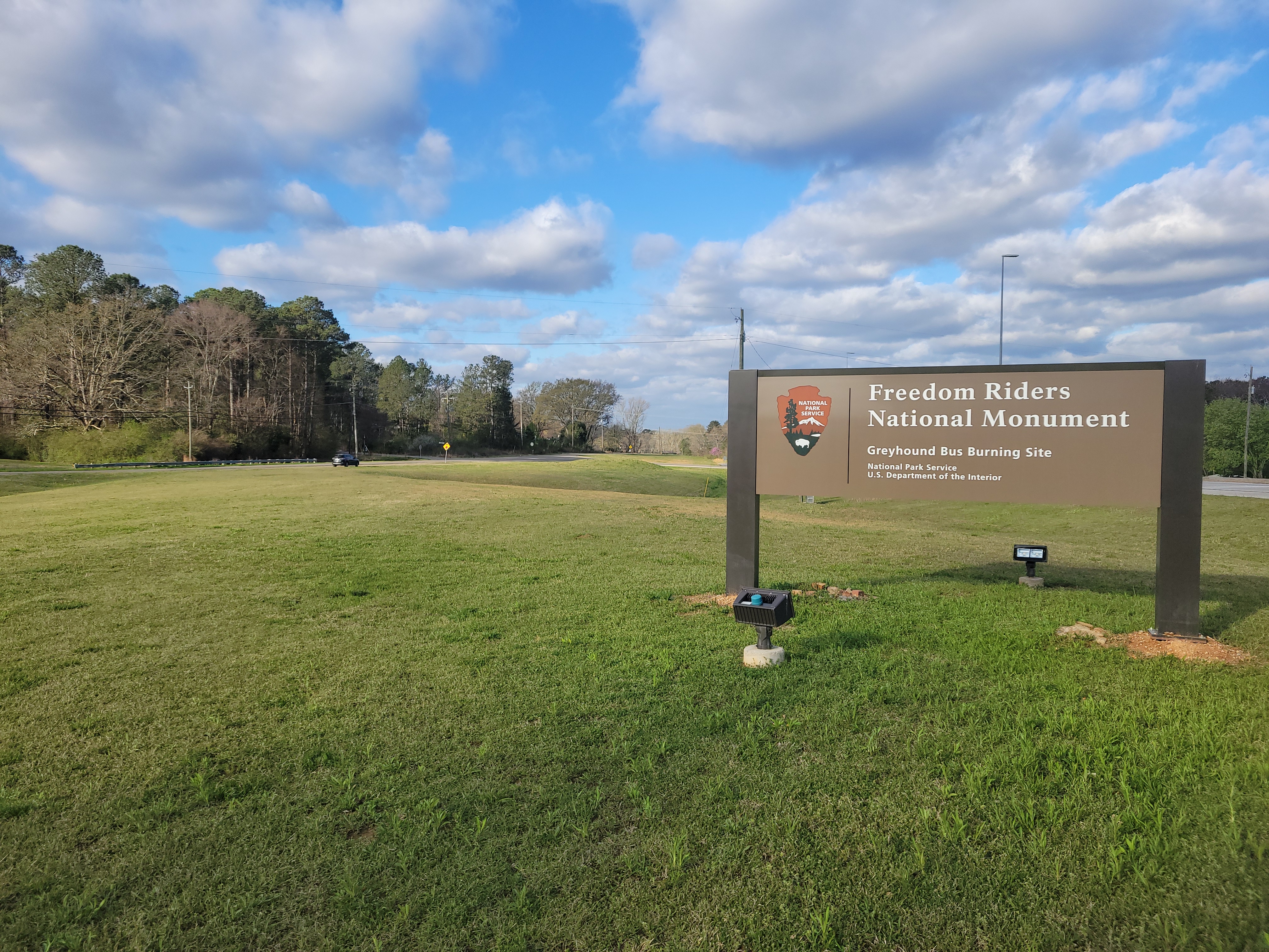 A sign saying Freedom Riders National Monument Bus Burning Site is posted in a grassy field.