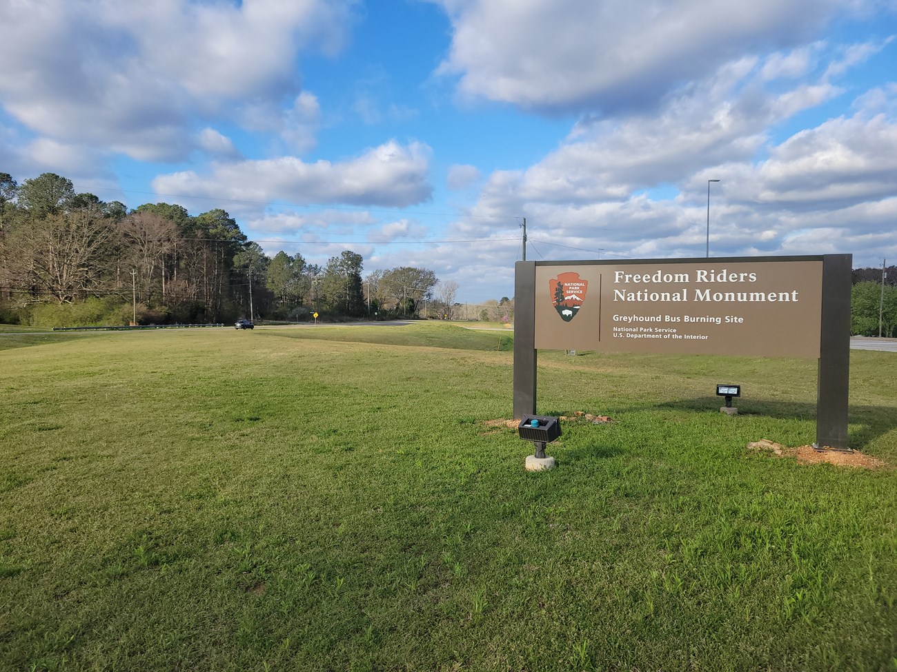 A sign saying Freedom Riders National Monument Bus Burning Site is posted in a grassy field.