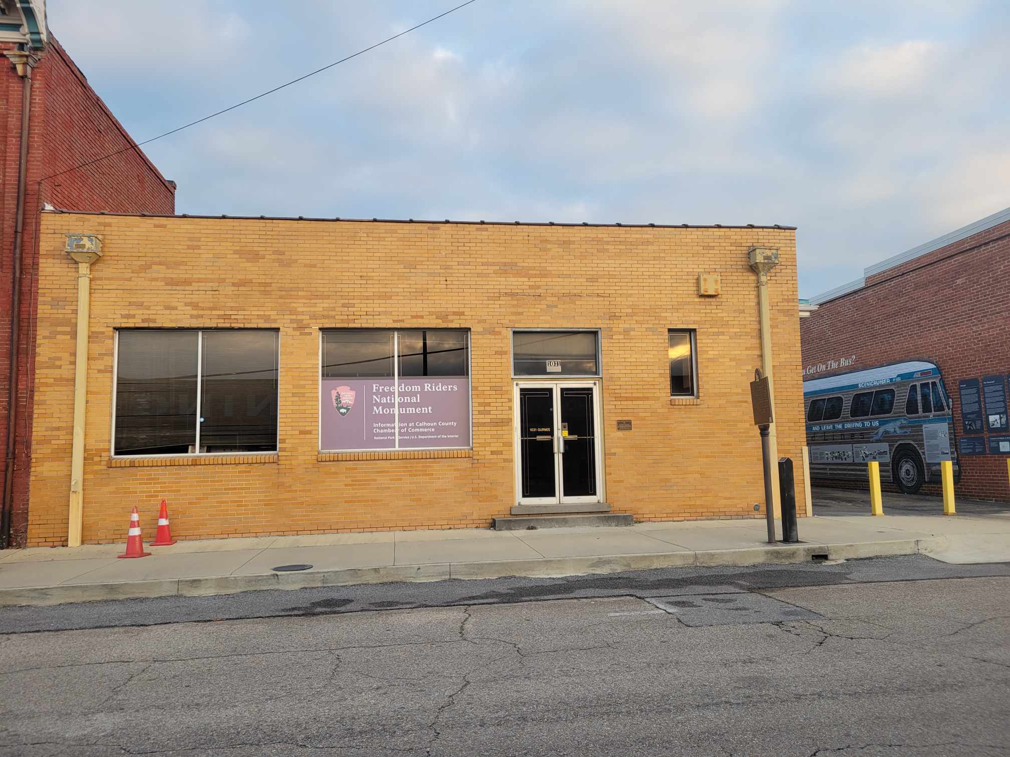 A brick building with large glass windows. A sign in a window says Freedom Riders National Monument. A mural is on a wall on the right side on the next building.