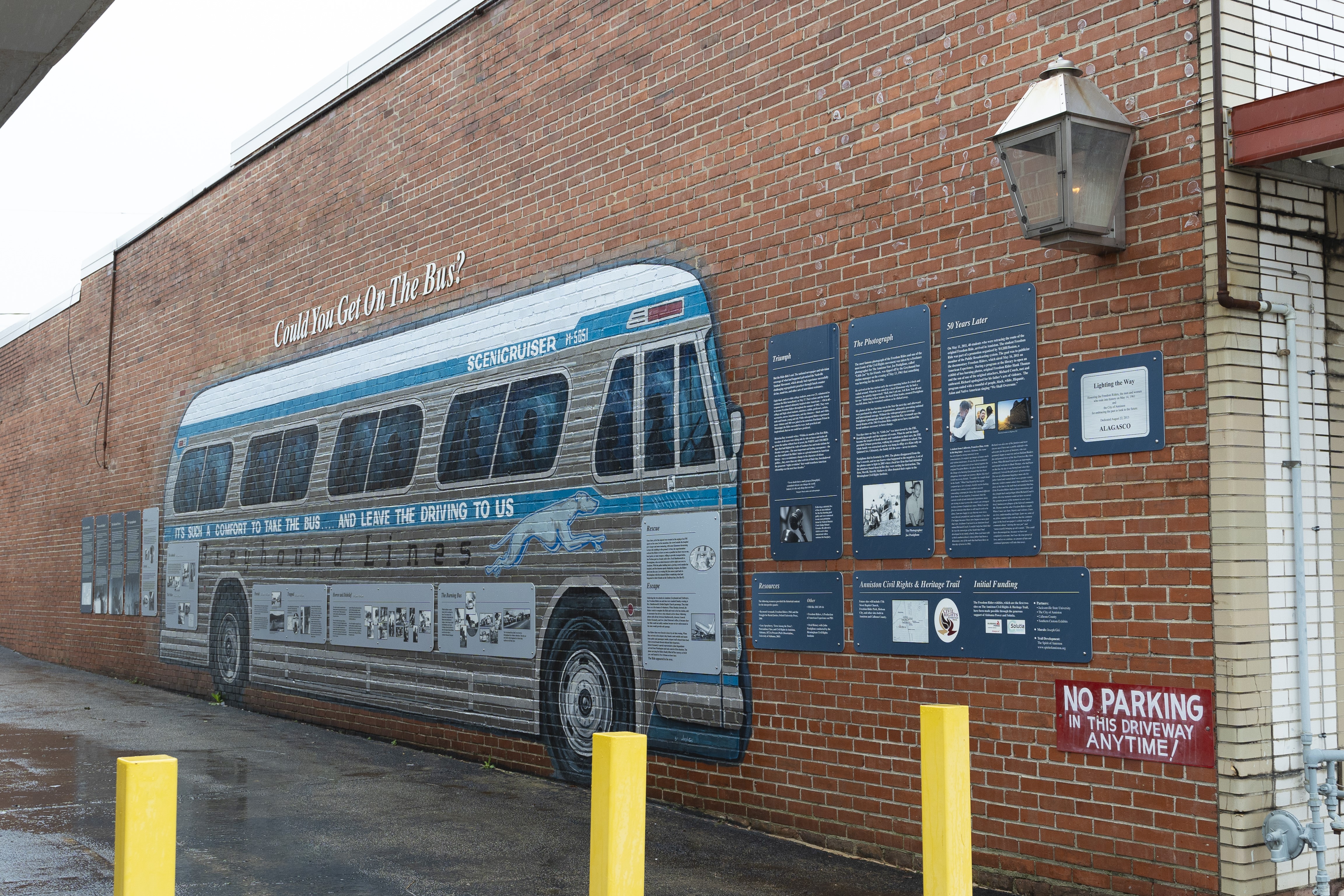 A mural of a bus is painted on a brick wall with the title "Could You Get On The Bus" above. Exhibit panels explaining the freedom rides are on either side and overlay the mural. A metal eternal flame is located high up on the wall on the right side.