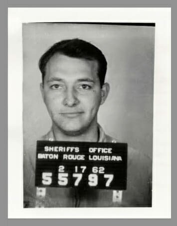A man is posing for a mug shot holding a sign that reads Sheriffs Office Baton Rouge Louisiana.