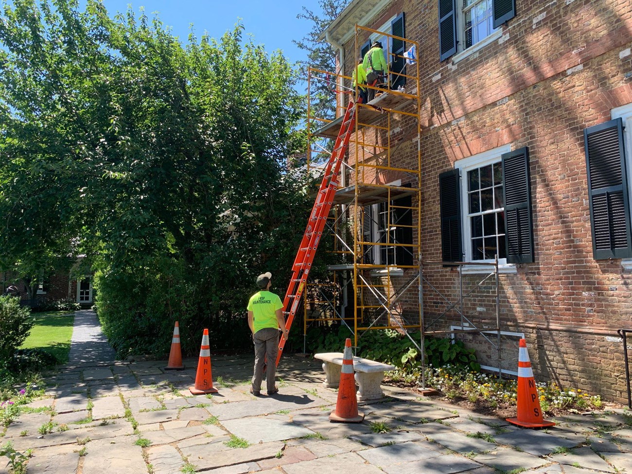 Two maintenance interns on a scaffolding platform near a historic building while another looks up on a ladder.