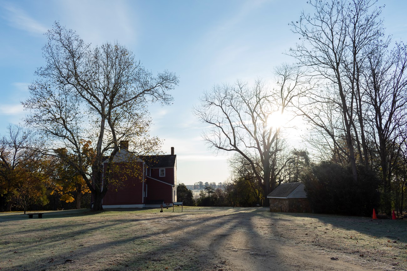 A gravel road leads to a two story red farmhouse surrounded by fields.