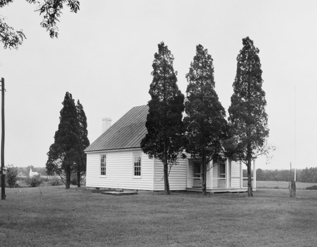 A white building in a cleared grassy field with tress obscuring the front door.