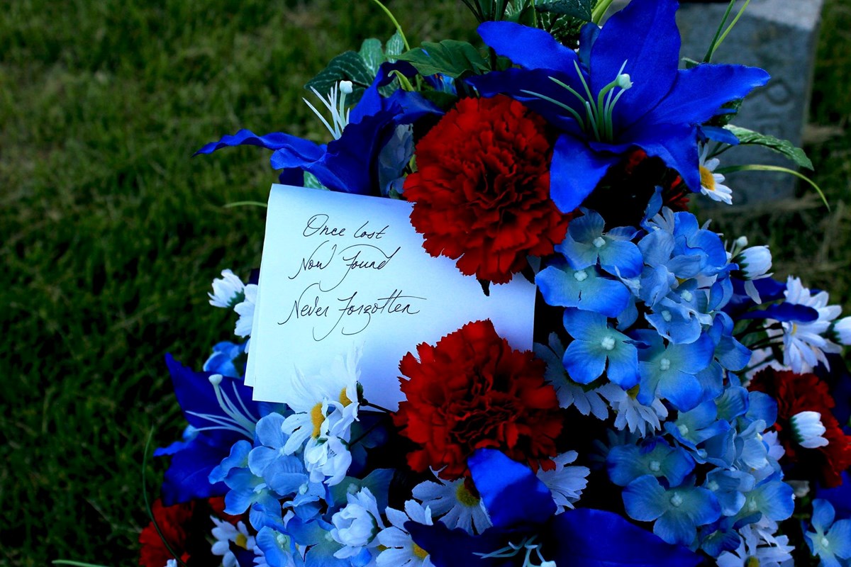 Red, white and blue flowers decorating grave with card reading "Once Lost, Now Found, Never Forgotten"