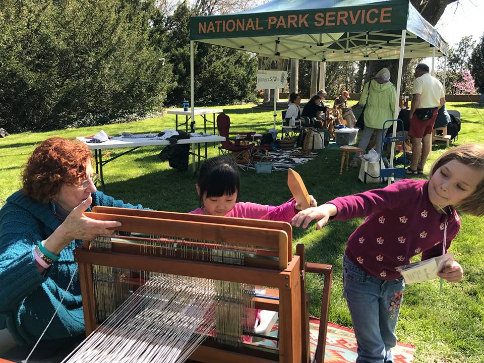 Two children using a loom while textile artisan observes, with green National Park Service tent in the background