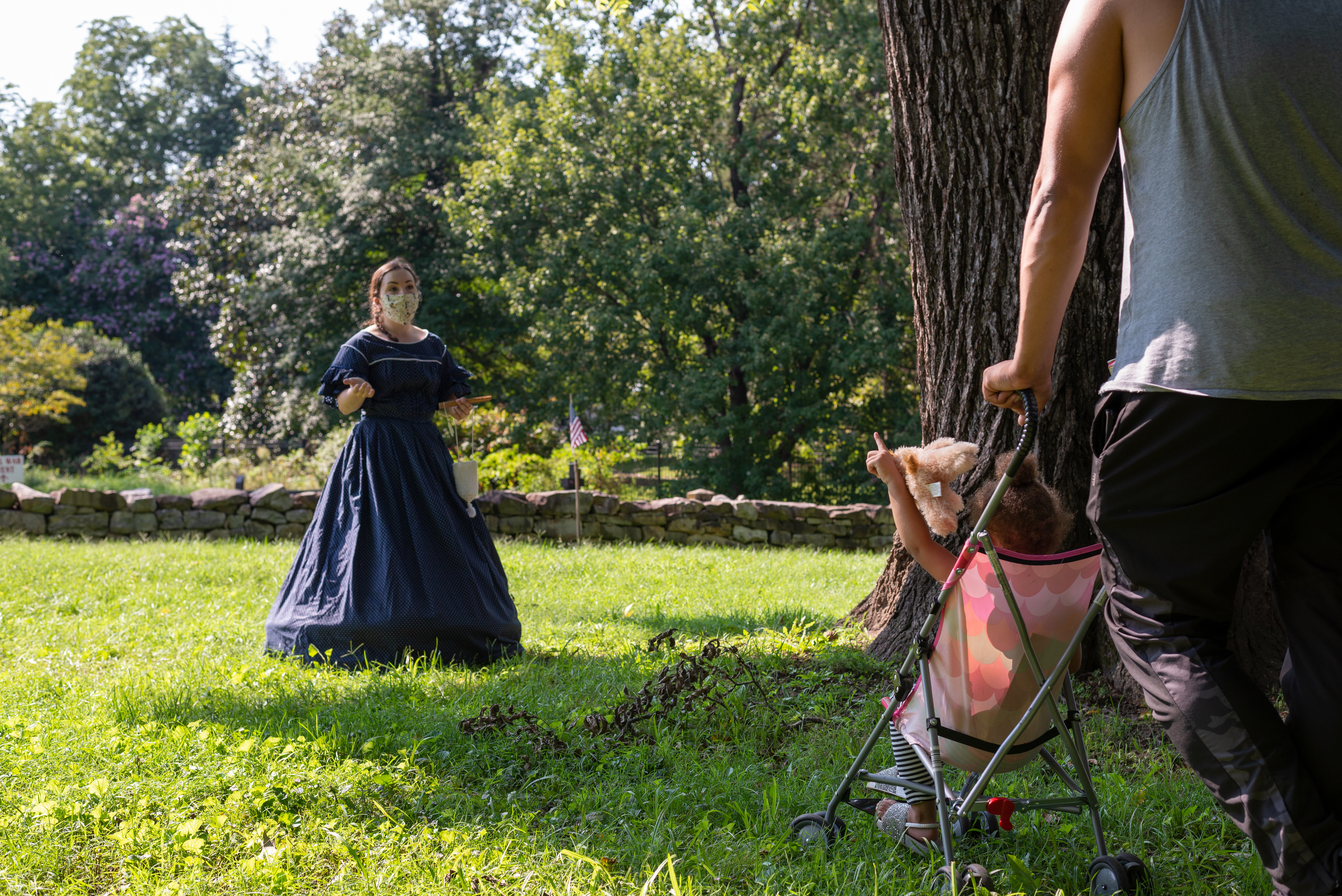 A woman in 1860s dress speaks to visitor as a child in a stroller raises her hand.