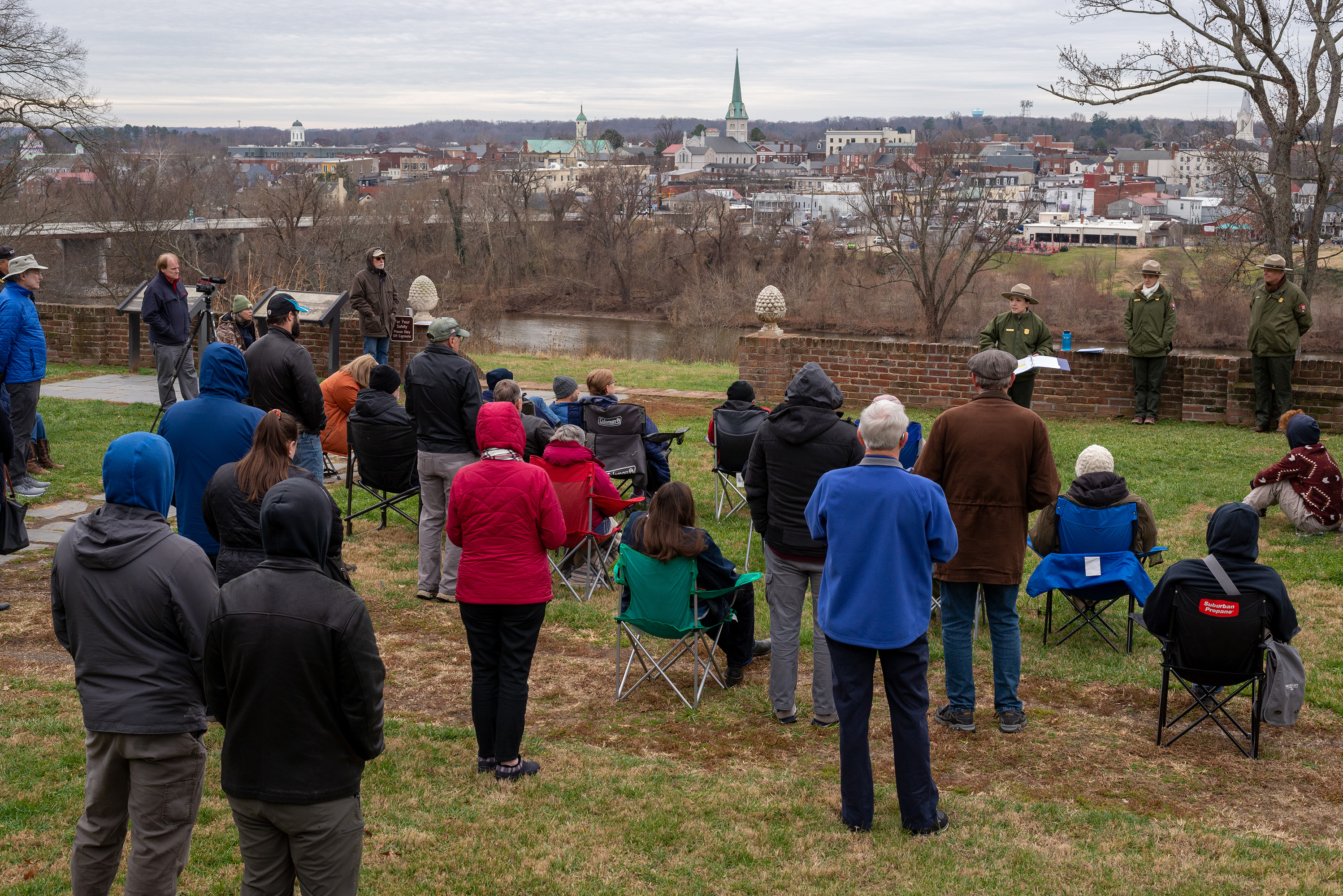 A female park ranger speaks to a group of 50 people on a lawn overlooking a river across from Frederickburg.