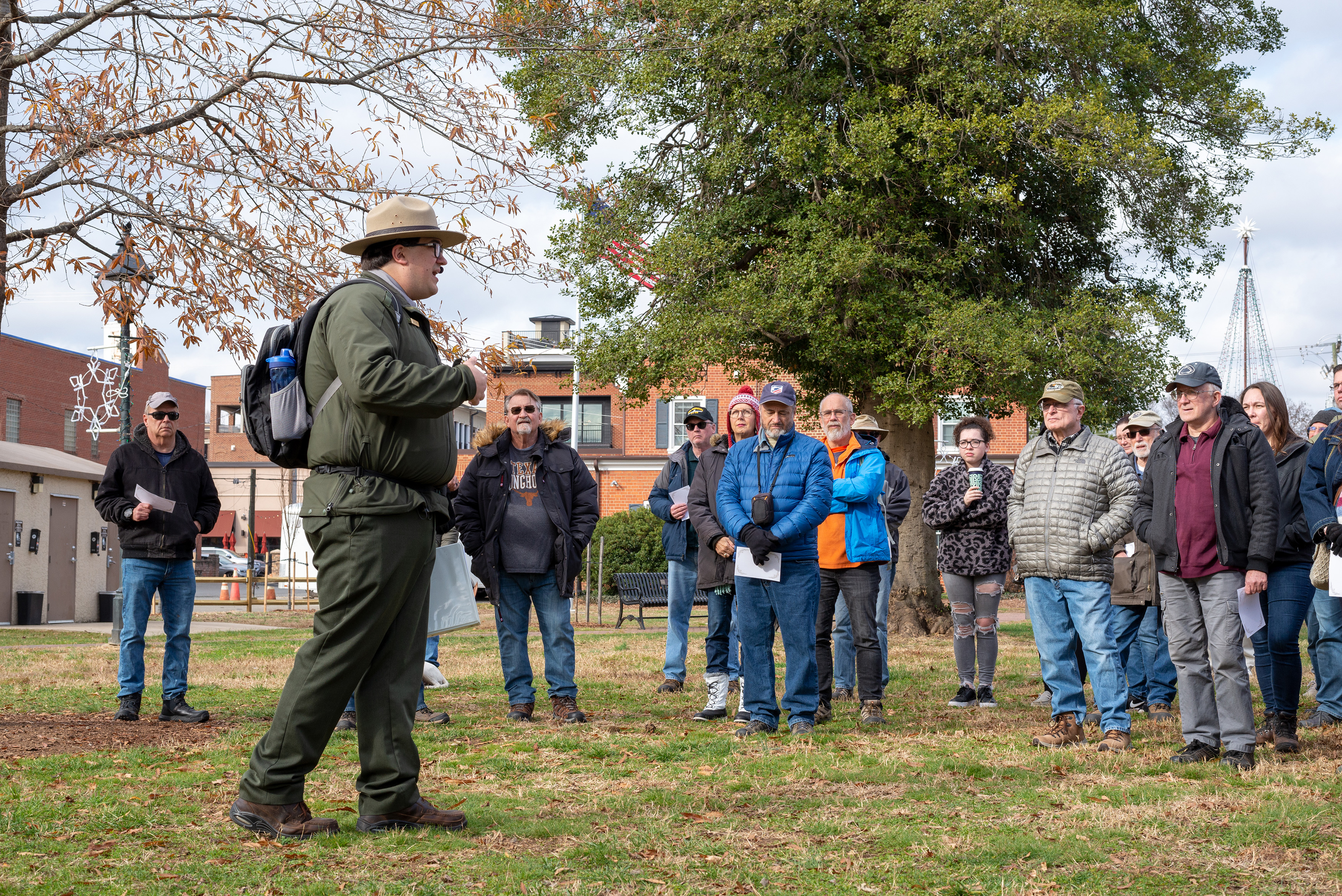 A male park ranger speak on the lawn of a city park to 20 people.