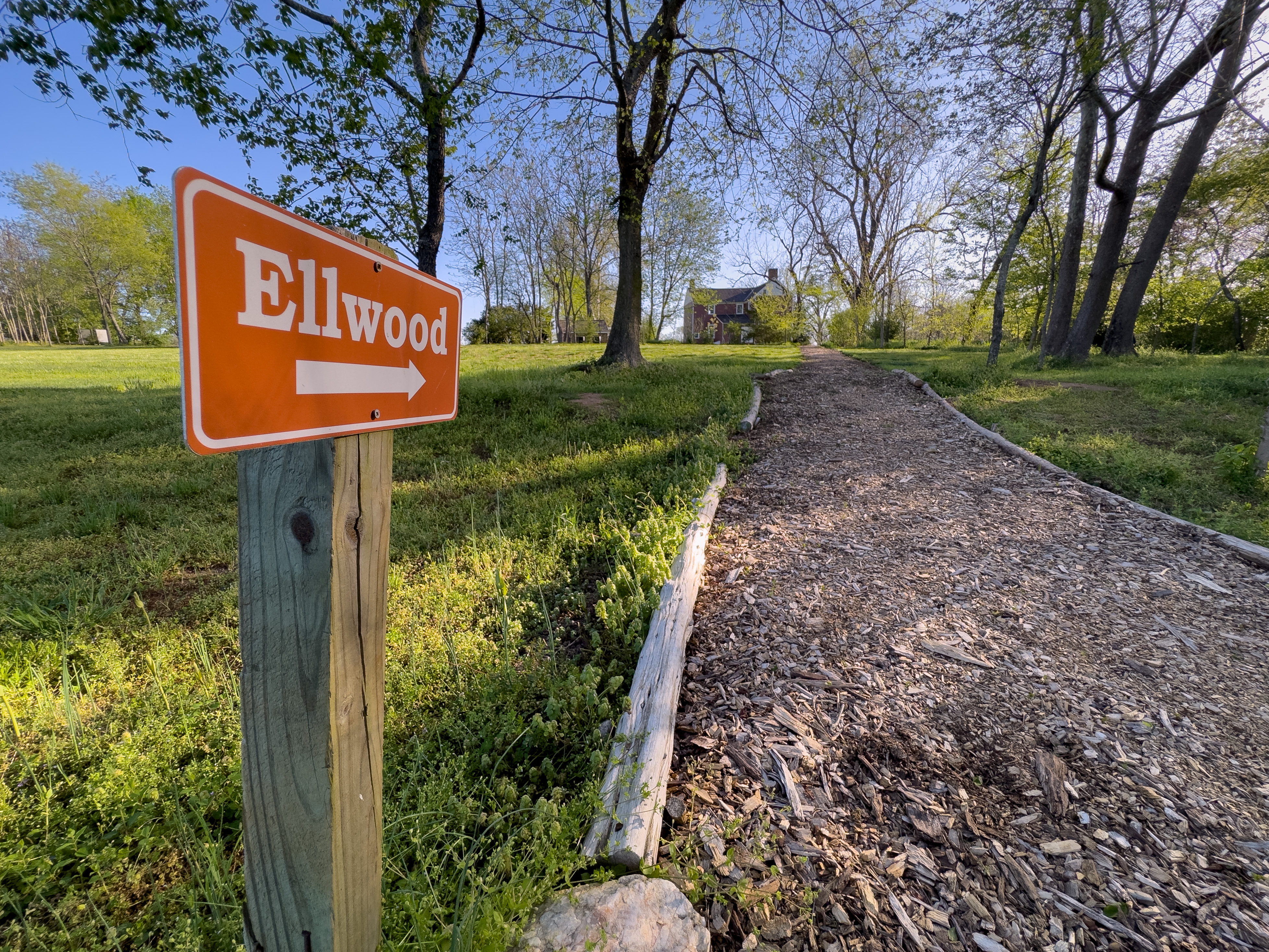 Ellwood in Spring, with sign A sign with the word Ellwood, pointed along a mulch path leading to a red farmhouse.