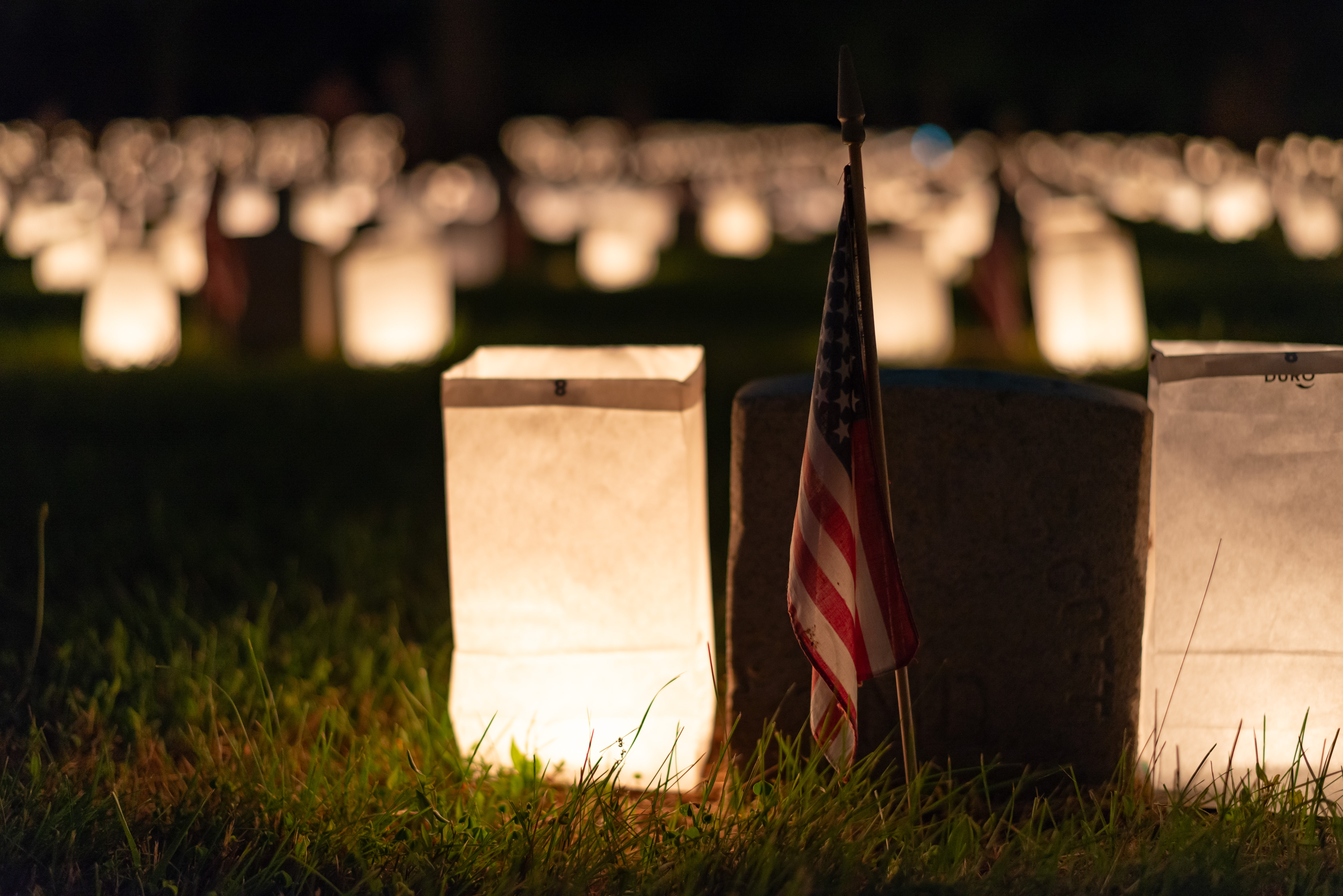 Luminaria 2024 Close Up A small American flag and two luminaria bags sit next to a grave stone in a dark cemetery.