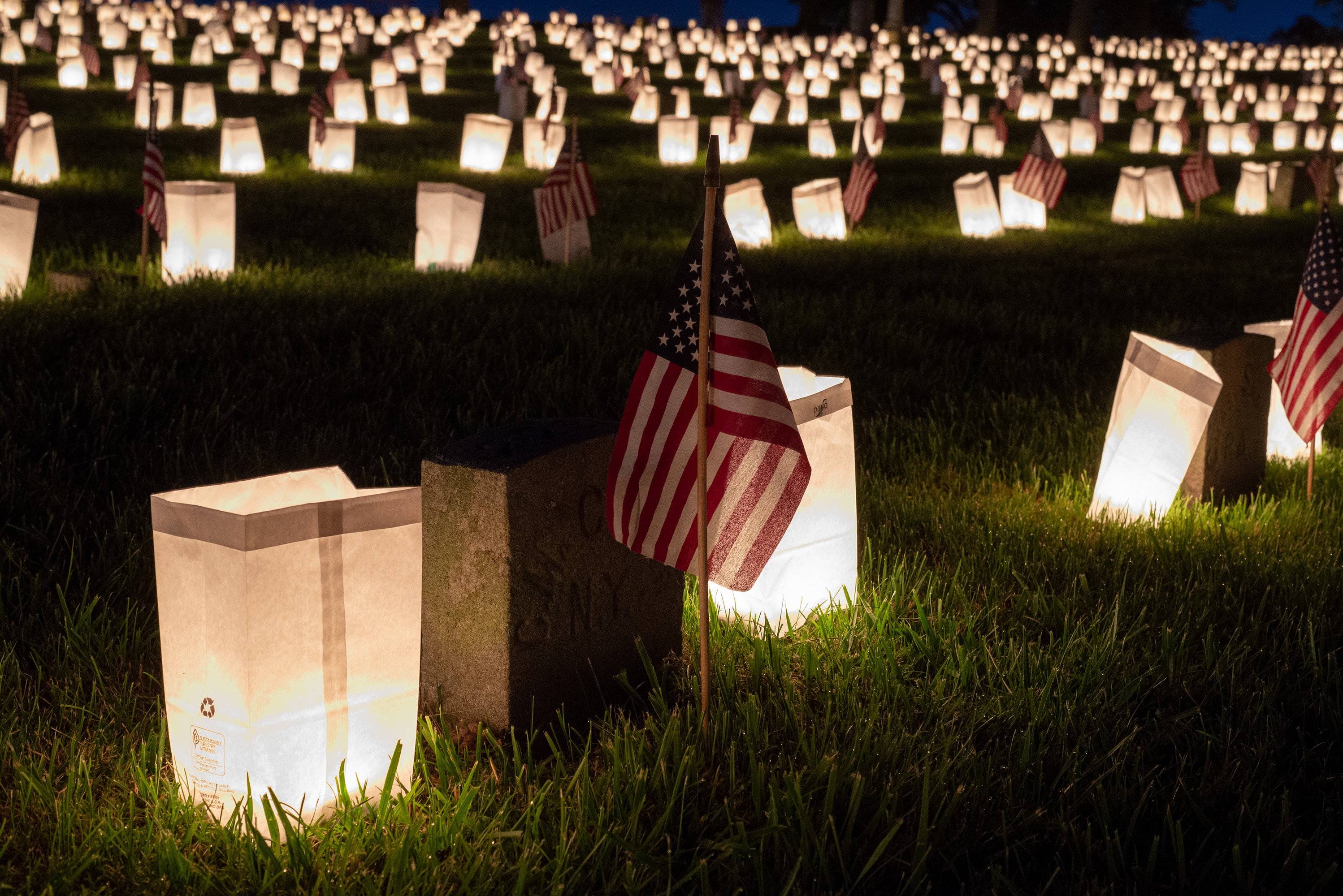 A headstone with two luminaria bags around it in a cemetery full of luminarias at night.