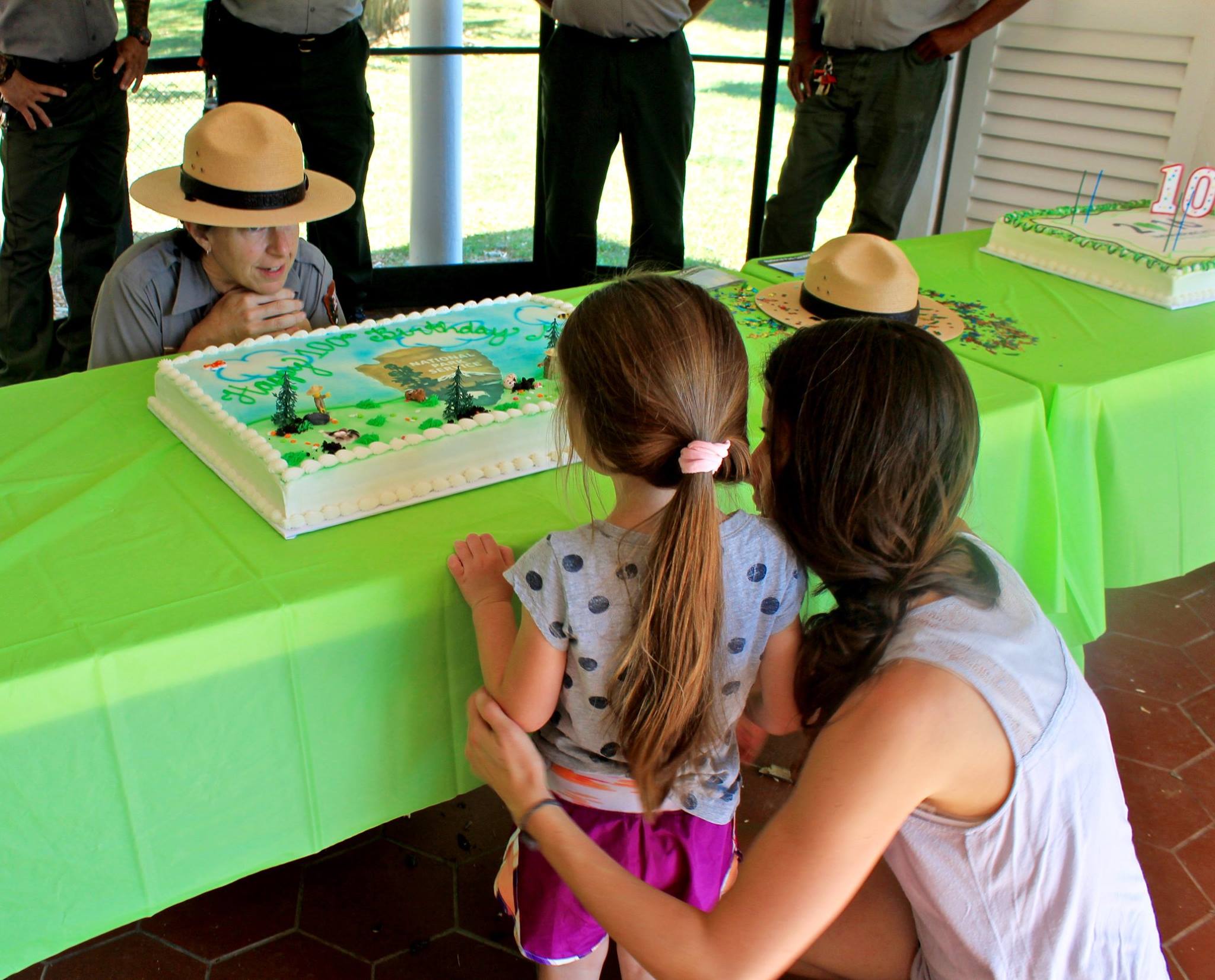 Park ranger with flat hat kneels behind table with cake to talk to small child and her mother