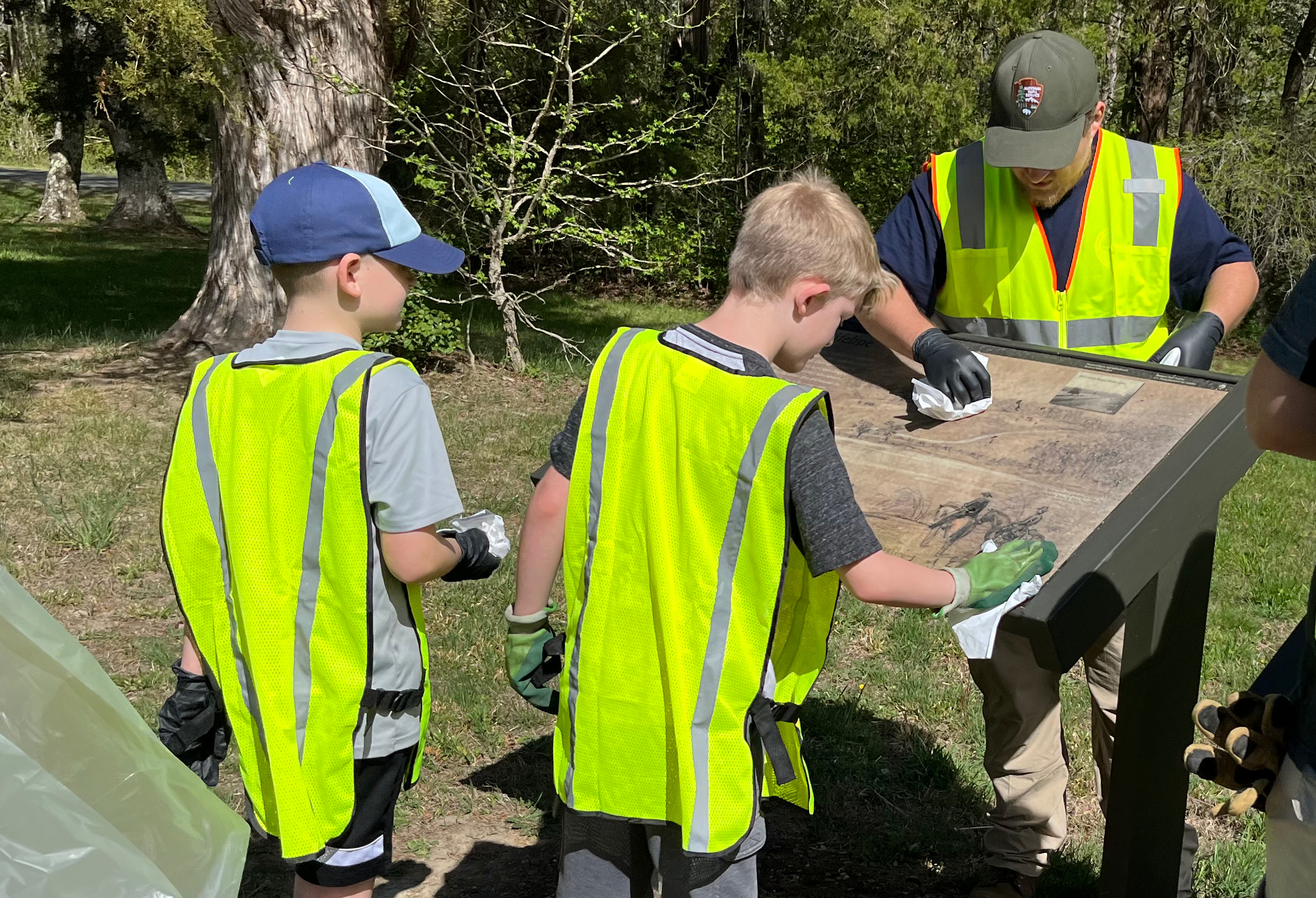 Two young boys clean a park sign with the guidance of a park staff member, all wearing safety vests.