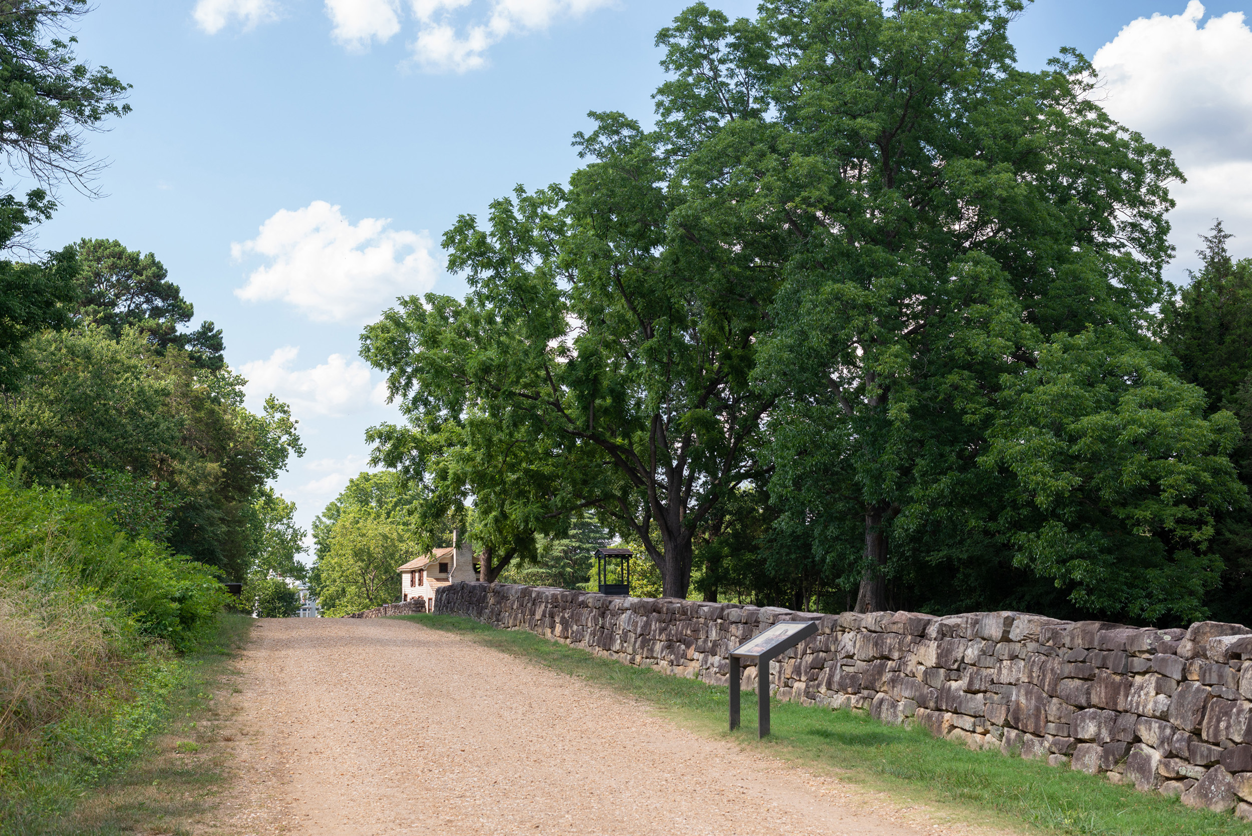 A gravel road bordered by a stone wall with a small, white house in the distance.