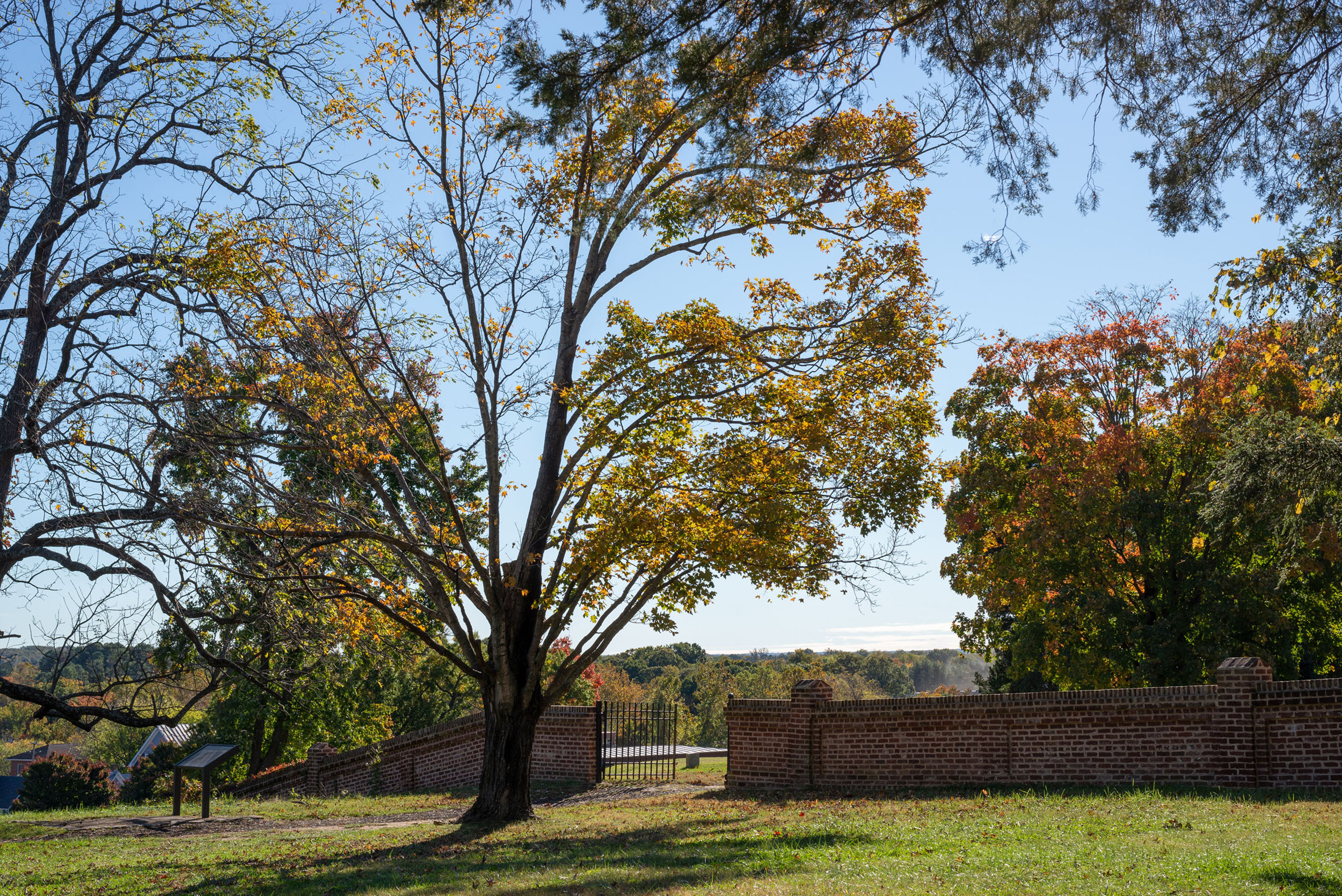 A view across a mowed field to a brick wall leading to a cemetery.