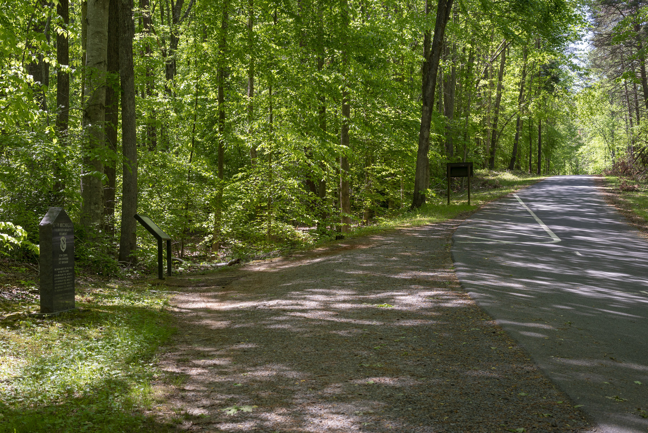 A small granite monument and interpretive sign and the side of a tree lined road.