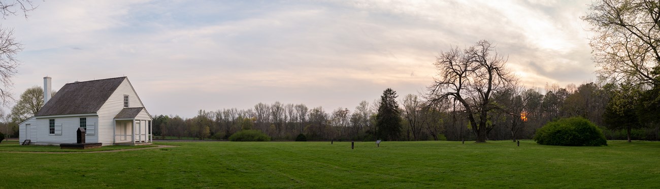 Panorama with small white structure on left and wide green field with sky stretching into a sunset on left.