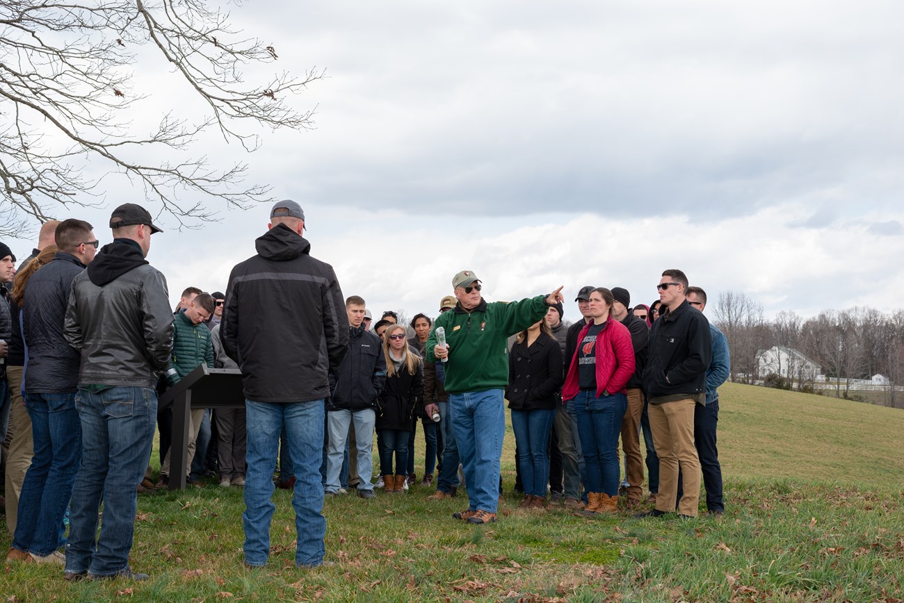 A National Park Service volunteer pointing to a field surrounded by 50 marines in civilian dress.