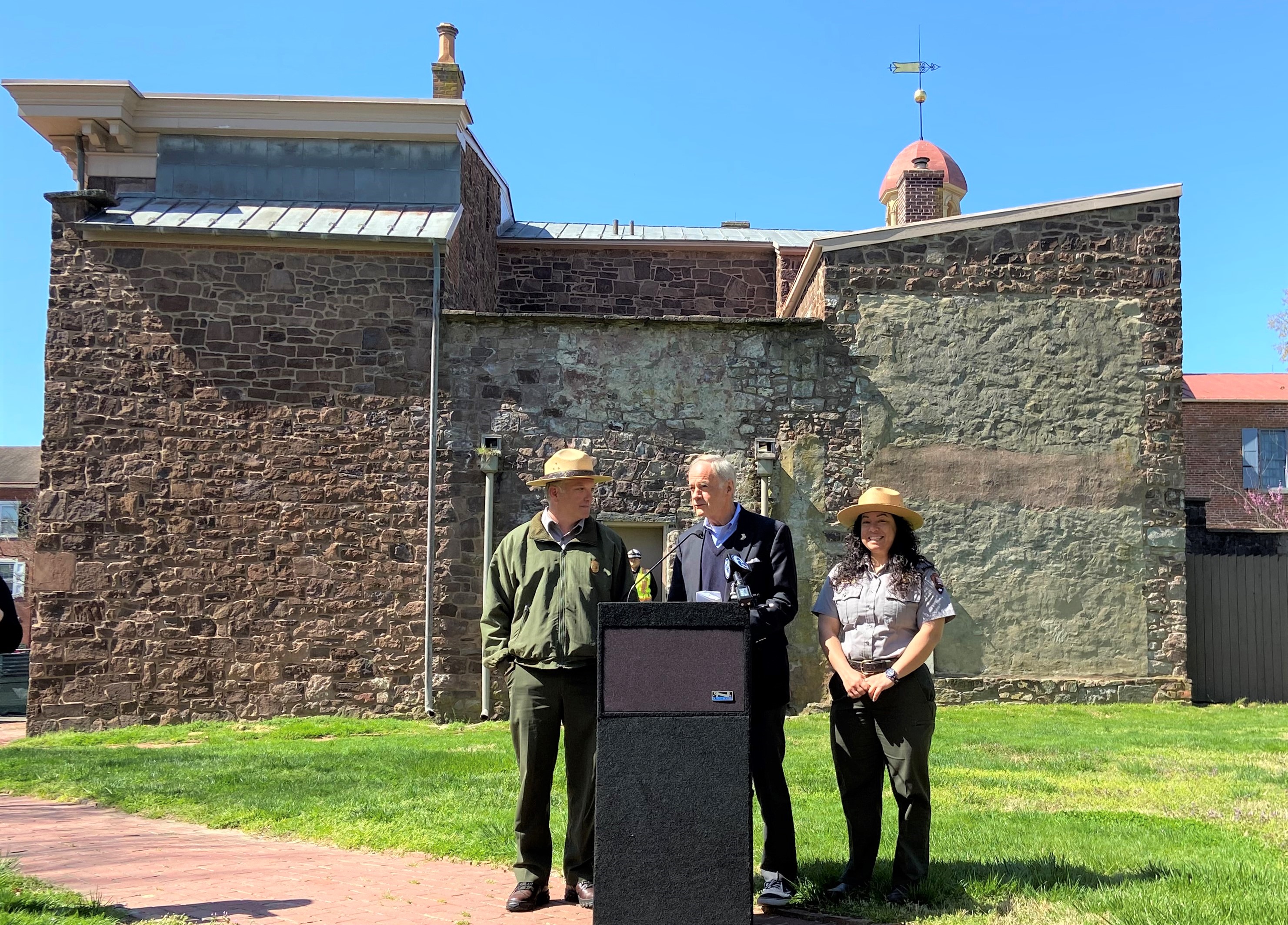 Two uniformed park rangers and another man stand behind a podium with a microphone.