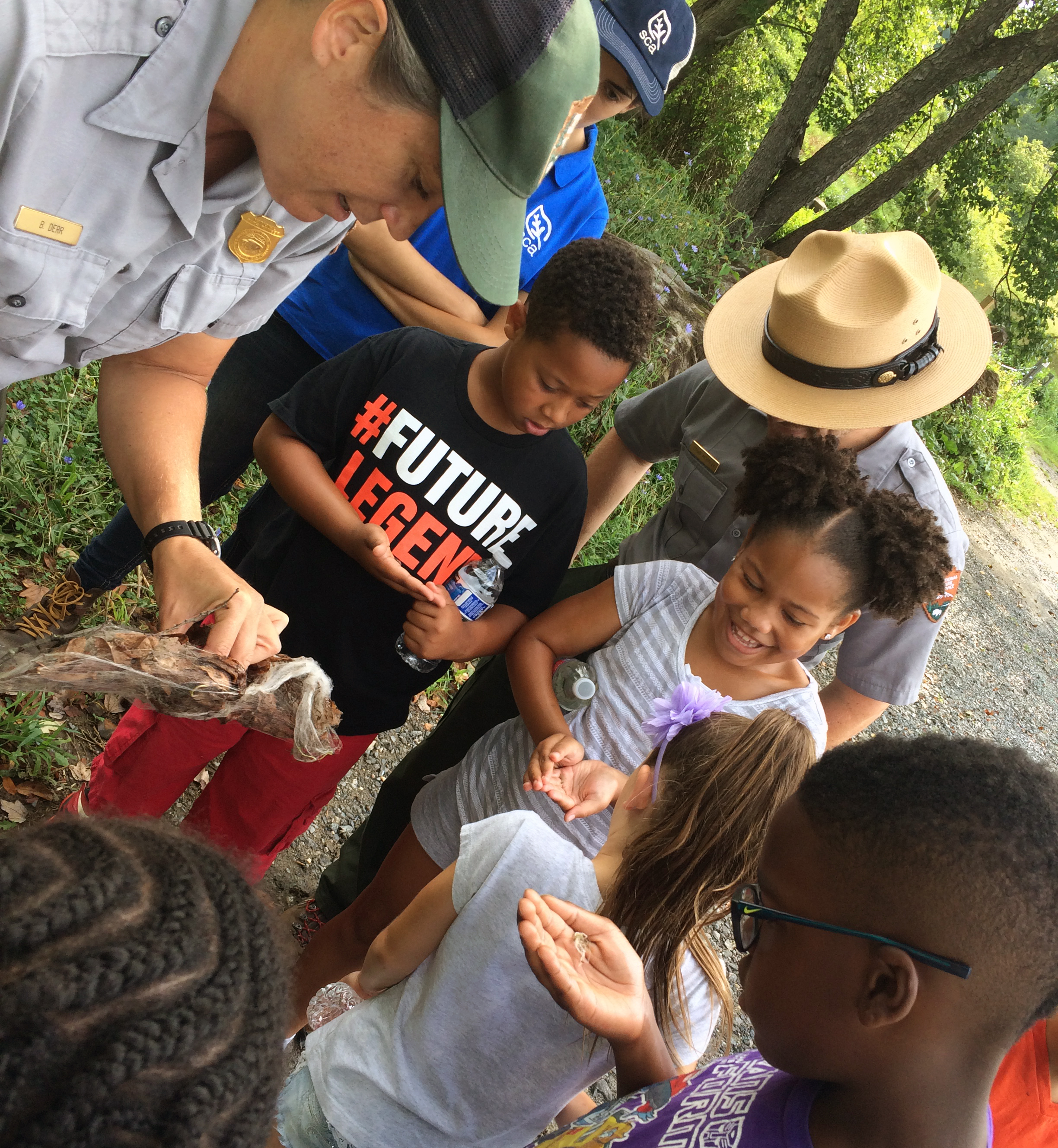 Children gather around a park ranger outdoors to look at insects.