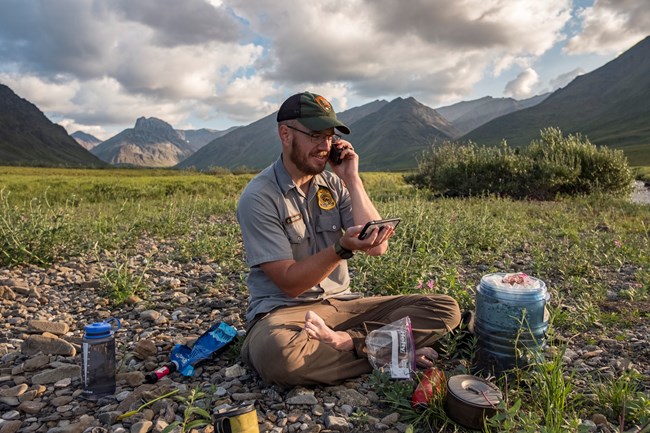 A park ranger calls in his GPS location to the Communication Center over a satellite phone while sitting on a gravel bar in the mountains