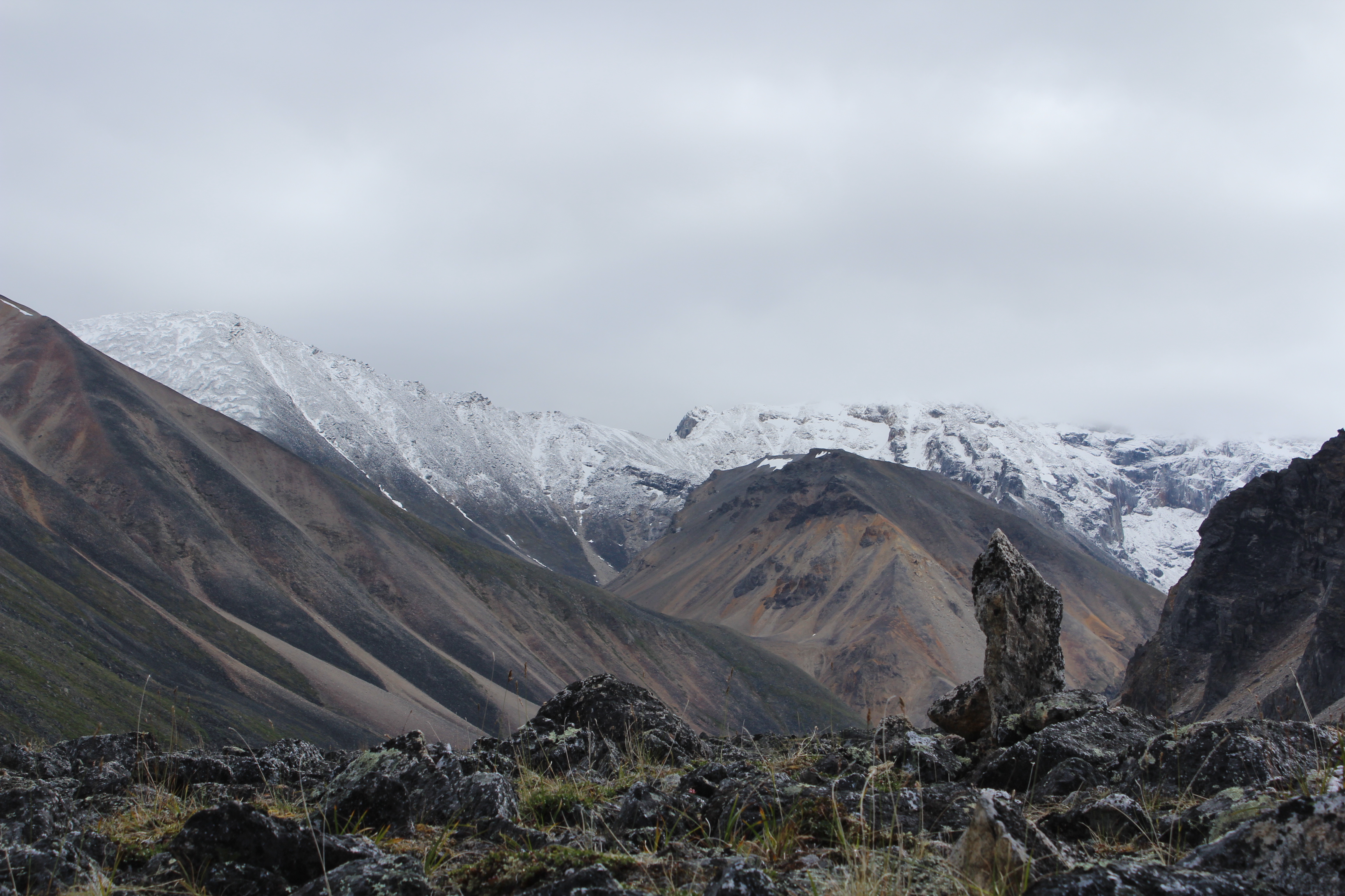 A solitary standing stone amid a lichen covered boulder field with snow covered peaks in the background.