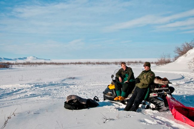 Two rangers sit and relax on a snowmachine on a sunny winter day