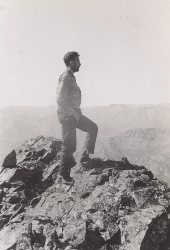 man stands on a top of a tall mountain, with other mountain peaks in the background