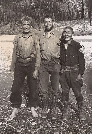 Three men stand together next to a river. The man in the middle has his arms on the shoulders of the other men