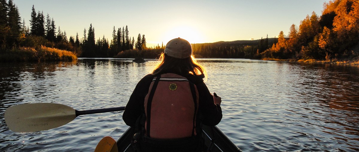 Woman paddling a canoe towards sunset on a forested river